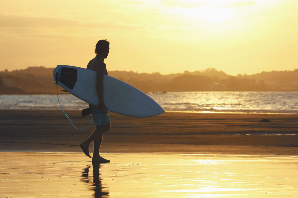A surfer walks towards the water in the afternoon sun